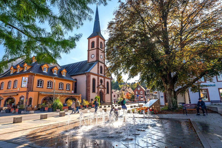 Place du centre de Niederbronn-les-Bains avec ses jeux d’eau, son église à clocher pointu et ses arbres