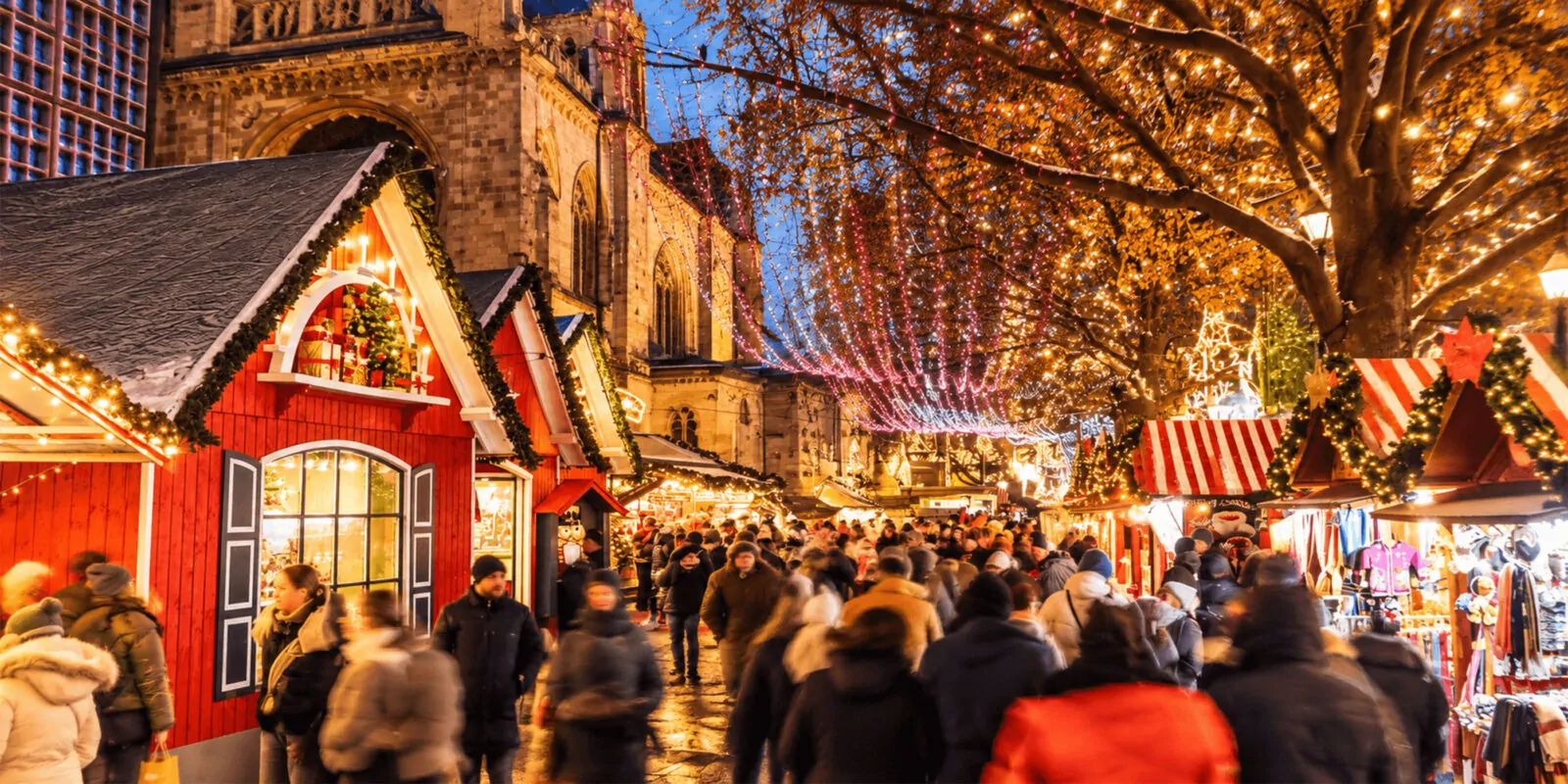 Marché de Noël illuminé à la tombée de la nuit, chalets décorés et foule sous des guirlandes lumineuses